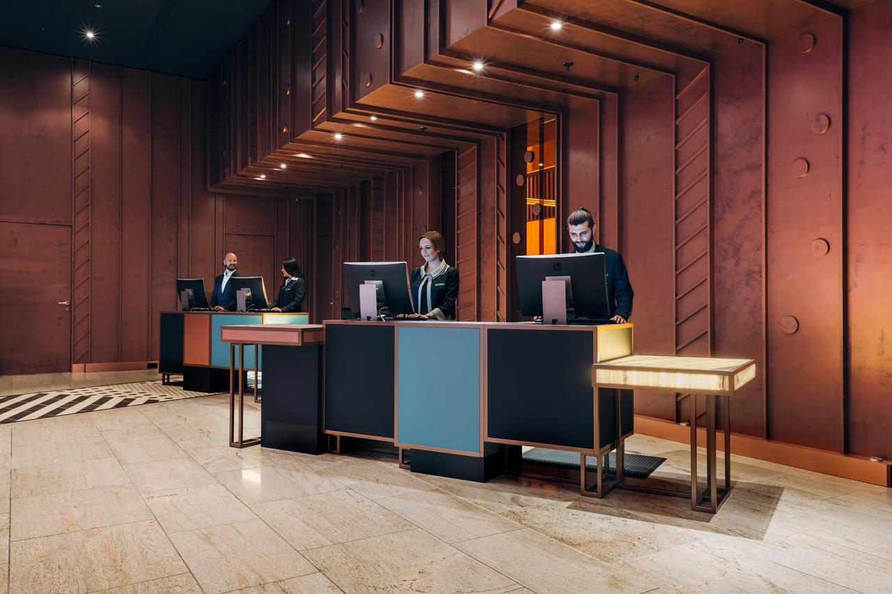 فندق بولمان برلين شفايتزرهوف المظهر الخارجي الصورة The image shows a modern hotel lobby reception area. There are several reception desks made of dark materials with illuminated accents. Behind the desks, three people are standing, likely staff members, engaged with computer screens. The lobby featur